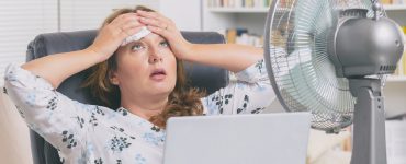 Frau schwitzt vor Hitze im Büro und kühlt sich mit einem Ventilator ab