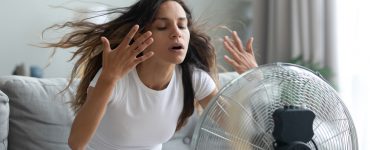 Eine junge Frau sitzt im Wohnzimmer vor einem Ventilator und versucht sich abzukühlen.