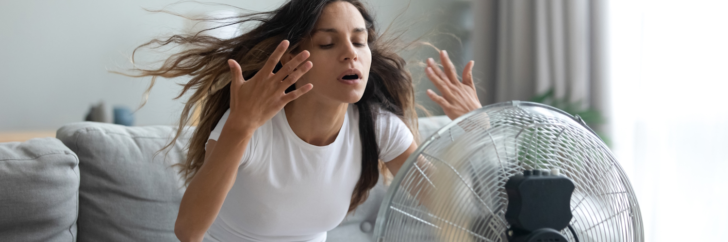 Eine junge Frau sitzt im Wohnzimmer vor einem Ventilator und versucht sich abzukühlen.
