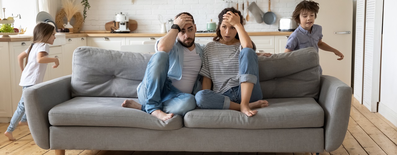 Müde Eltern sitzen auf der Couch und Kinder rennen durch die Wohnung