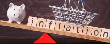 Ein Holzbrett als Waage. Auf der linken Seite auf der Waage ein Sparschwein, auf der Rechten Seite das Wort Inflation