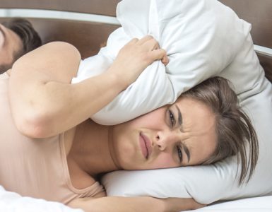 Eine Frau liegt neben einem schnarchendem Mann im Bett und drückt sich die Ohren mit dem Kissen zu.