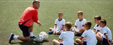 Eine Kinderfußballgruppe sitzt auf dem Rasen. Vor ihnen kniet ein Trainer. Vor dem Trainer liegt ein Fußball.