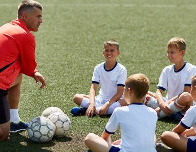 Eine Kinderfußballgruppe sitzt auf dem Rasen. Vor ihnen kniet ein Trainer. Vor dem Trainer liegt ein Fußball.
