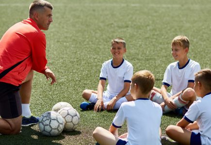 Eine Kinderfußballgruppe sitzt auf dem Rasen. Vor ihnen kniet ein Trainer. Vor dem Trainer liegt ein Fußball.