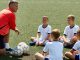 Eine Kinderfußballgruppe sitzt auf dem Rasen. Vor ihnen kniet ein Trainer. Vor dem Trainer liegt ein Fußball.