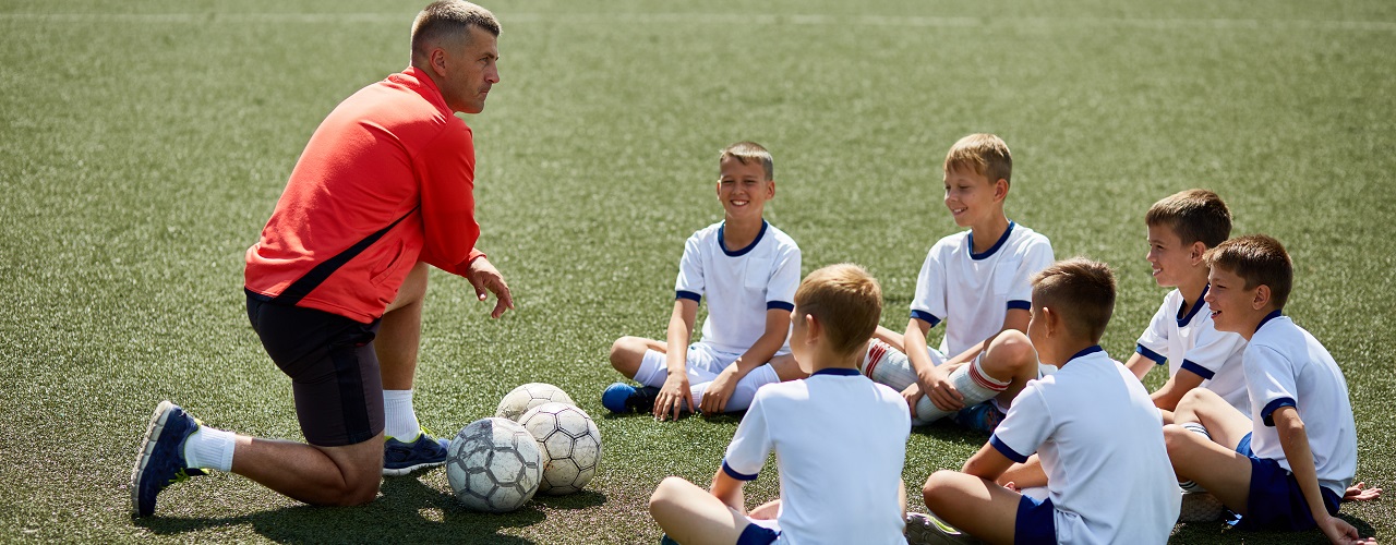 Eine Kinderfußballgruppe sitzt auf dem Rasen. Vor ihnen kniet ein Trainer. Vor dem Trainer liegt ein Fußball.