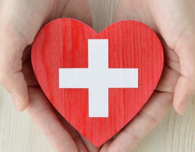 In der Mitte ein rotes Herz aus Holz. darauf befindet sich ein weißes Kreuz. Das Herz wird in zwei Händen gehalten. Am linken Rand liegt ein Stethoskop.