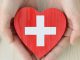 In der Mitte ein rotes Herz aus Holz. darauf befindet sich ein weißes Kreuz. Das Herz wird in zwei Händen gehalten. Am linken Rand liegt ein Stethoskop.
